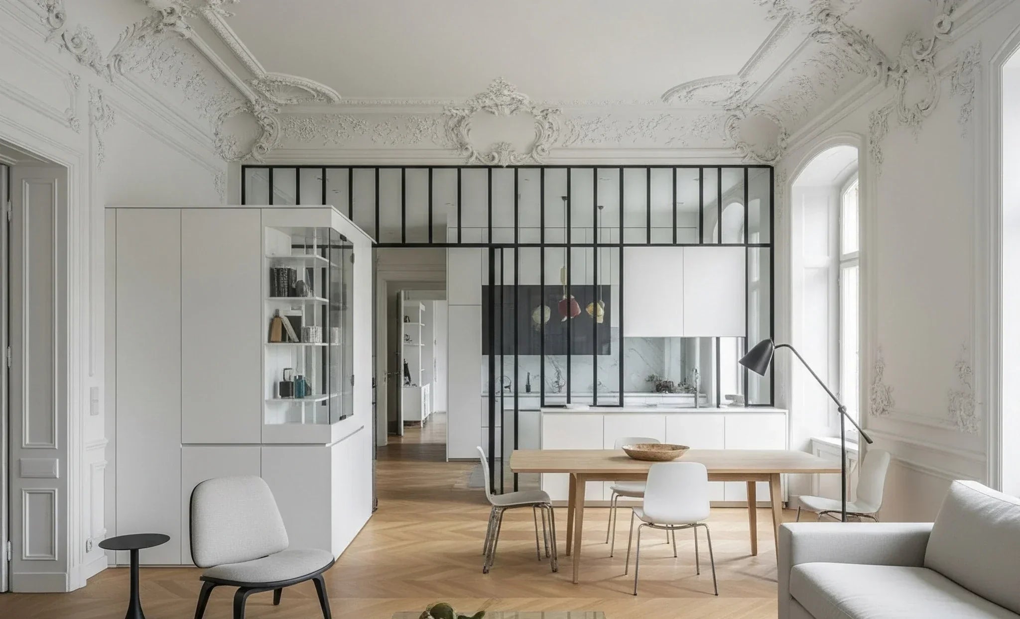 White room with ornate Rococo plasterwork, modern white furniture, light wood floor, and black-framed glass partition leading to a kitchen
