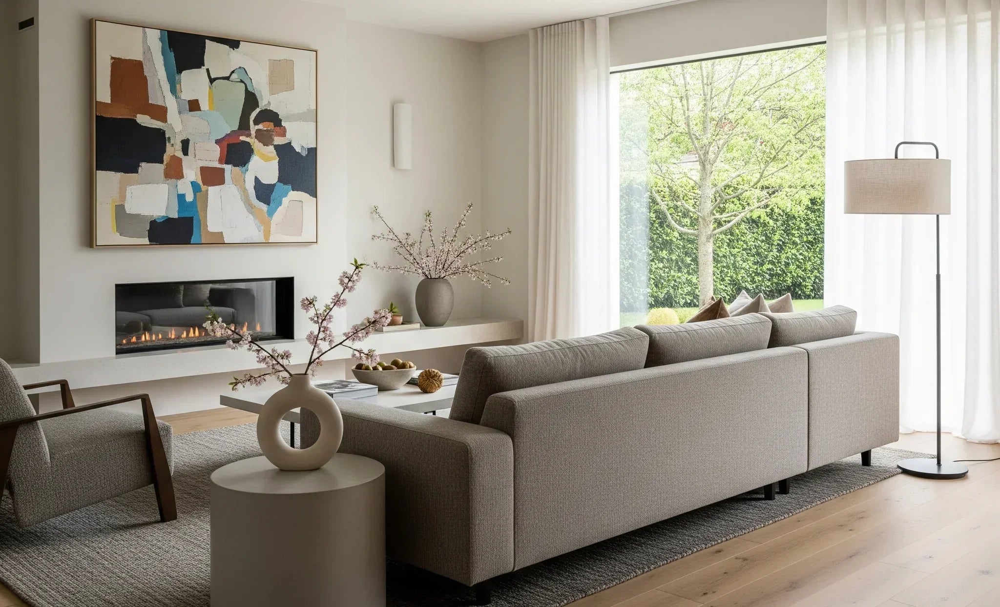 A living room with a large gray sofa, a fireplace, a large abstract painting, and a wide window with sheer curtains.