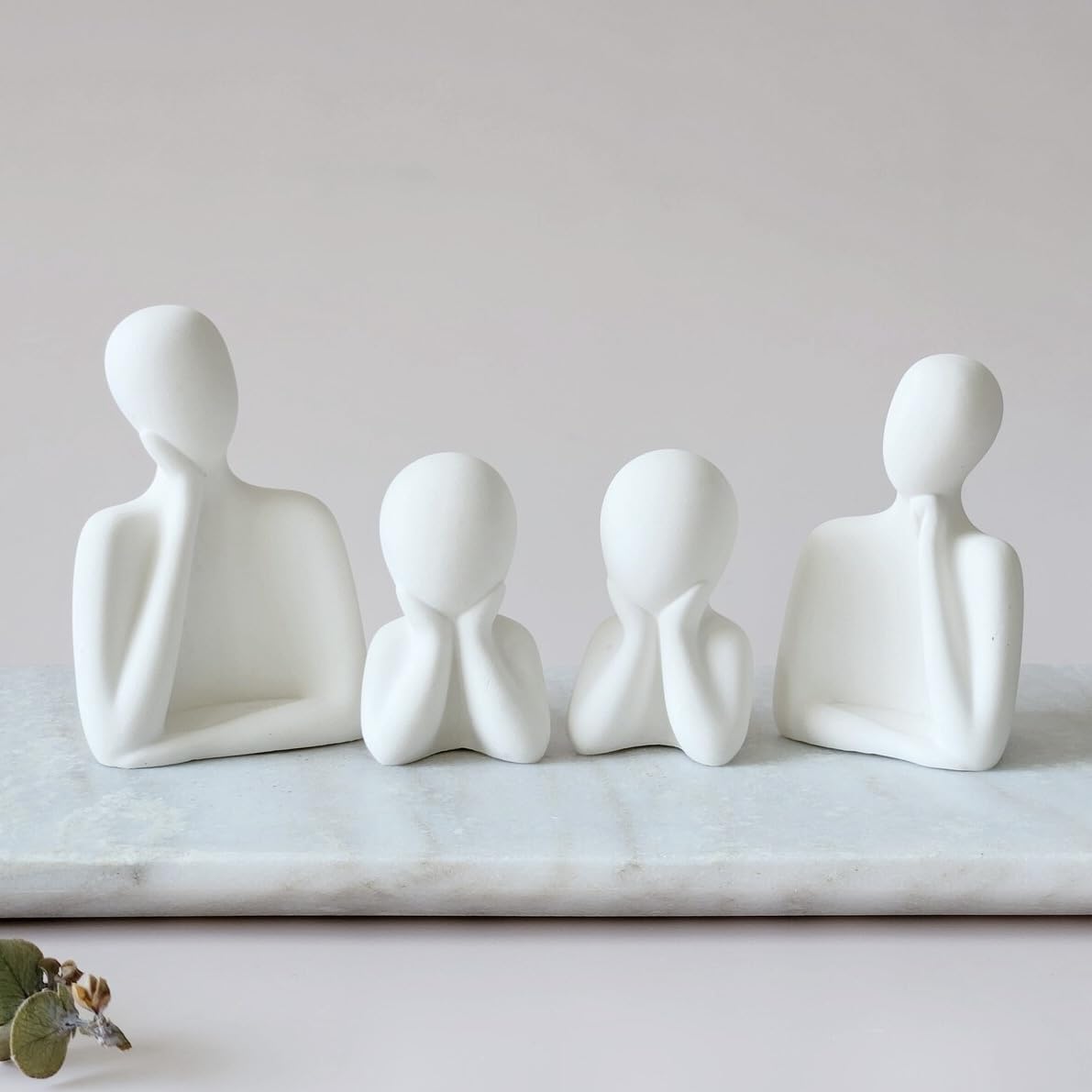 Four white abstract human figure sculptures on a marble surface with a plain background
