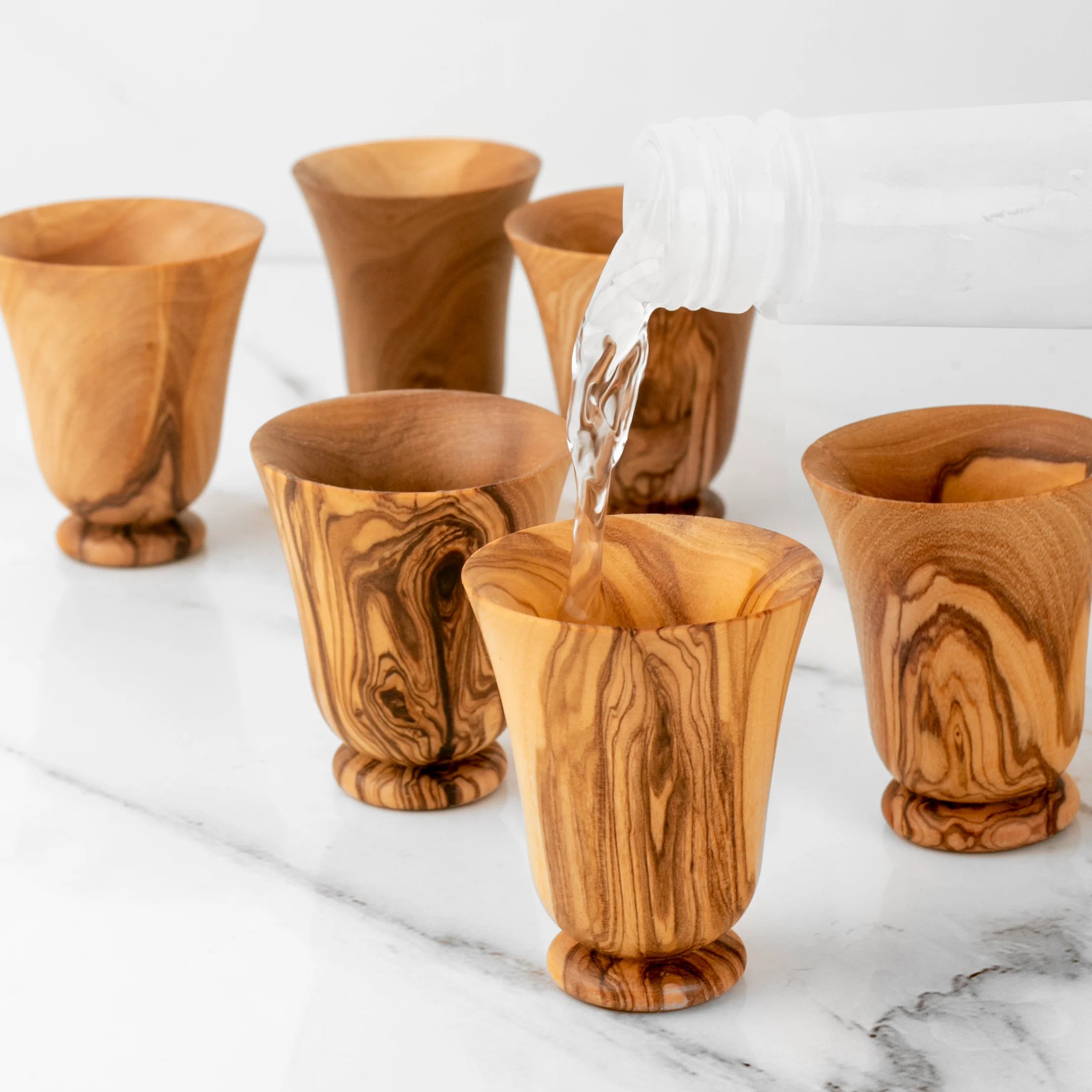 Wooden tumblers on a marble surface with one being filled with a clear liquid.