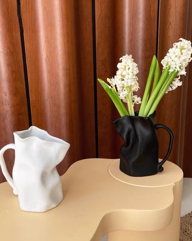Two pitchers, one white and one black, with white flowers on a wooden surface against a brown curtain.