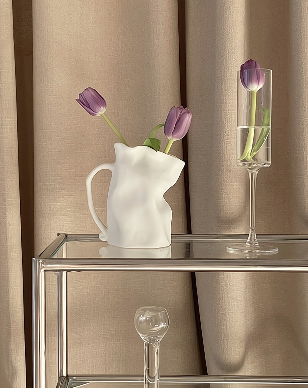 White pitcher with purple tulips on a glass table against beige curtains