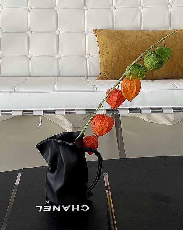 Black vase with red and green fruits on a black surface with 'Chanel' branding, against a white textured wall.
